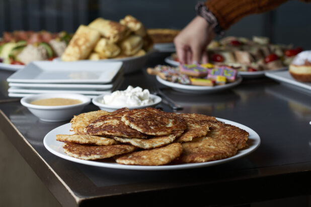 A plate of latkes on a buffet table of Hanukkah food with a hand reaching for cookies in the background
