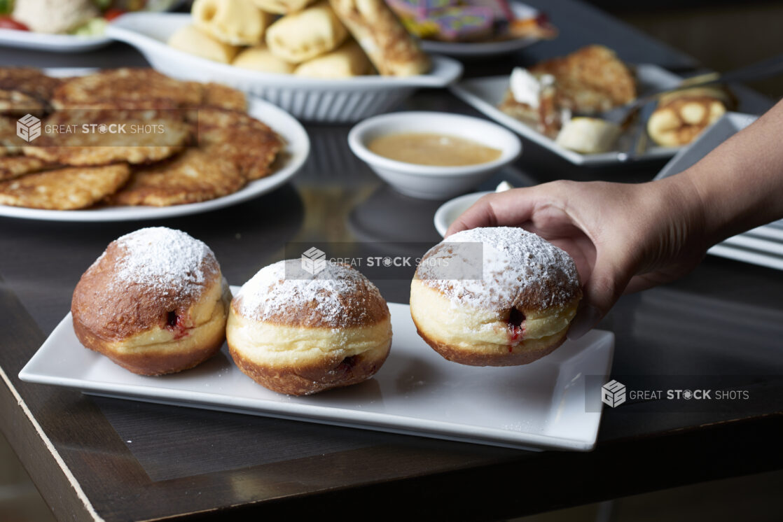 A hand picking up a sufganiyah from a buffet of assorted plates of Hanukkah food