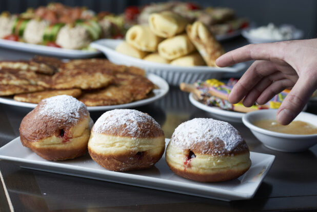 A hand reaching towards a sufganiyah on a buffet with assorted plates for Hanukkah