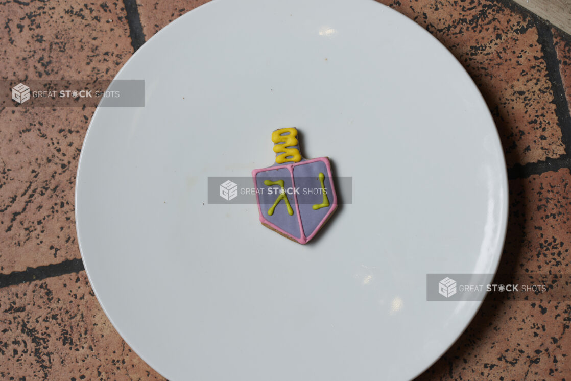 A decorative blue and yellow dreidel cookie on a white plate on a tile background