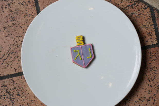 A decorative blue and yellow dreidel cookie on a white plate on a tile background