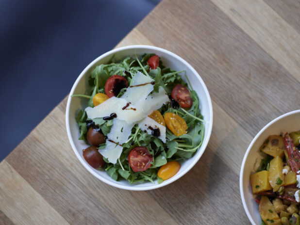 Arugula salad with shaved Parmesan and cherry tomatoes in a white bowl, other dish to the side