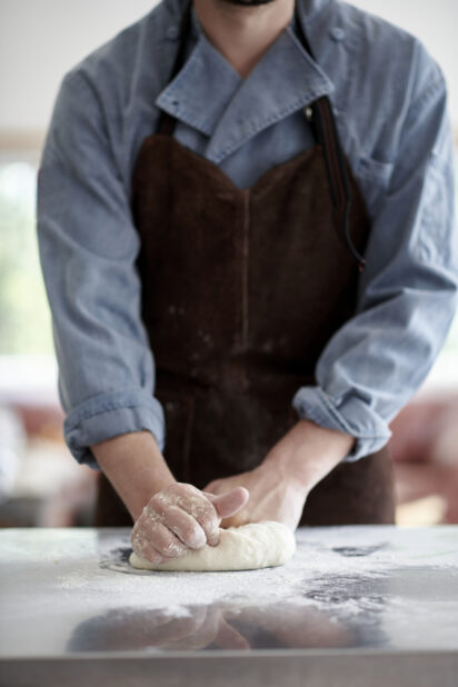 A chef in a dark apron kneading dough in flour on a steel prep table