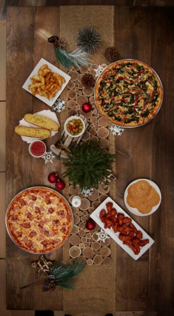 A dark wood dinner table decorated for Christmas with two large pizzas, a platter of wings, garlic bread, potato wedges and cookies, overhead view