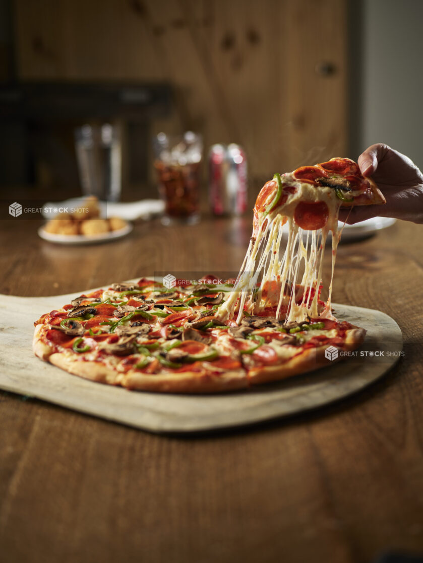 A hand lifting a slice from a traditional style pepperoni green pepper and mushroom pizza with chicken nuggets and a soft drink in the background