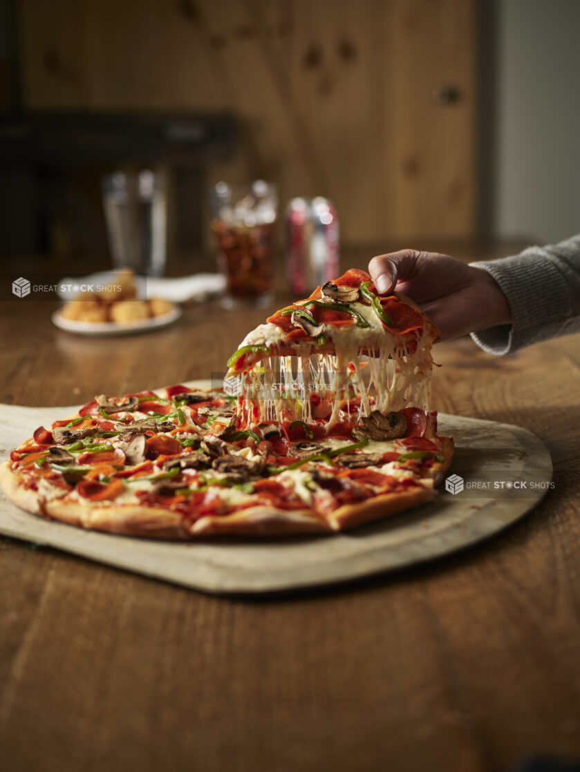 A hand lifting a slice from a traditional style pepperoni green pepper and mushroom pizza with chicken nuggets and a soft drink in the background