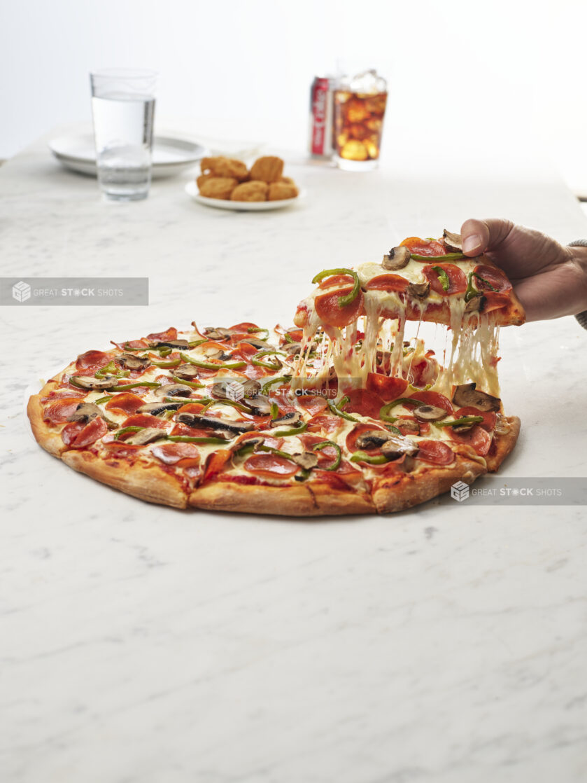 A hand lifting a slice from a traditional style pepperoni green pepper and mushroom pizza with chicken nuggets and a soft drink in the background