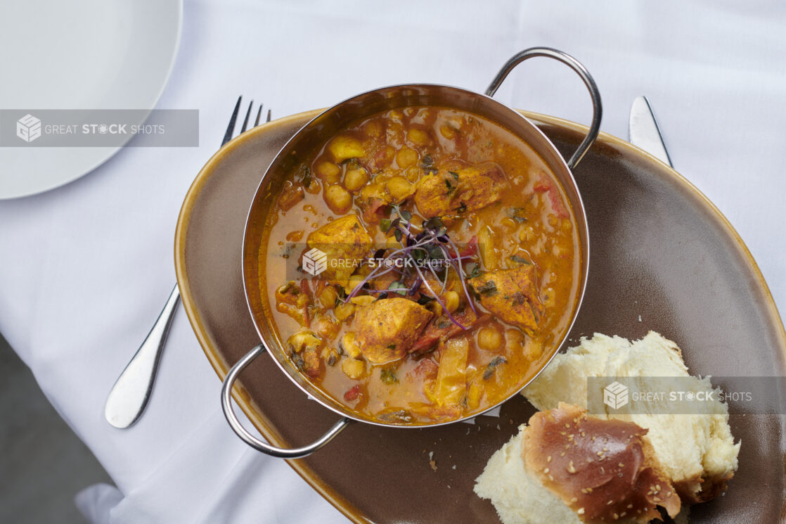 Butter chicken in a metal karahi bowl with bread on the side on a rustic ceramic plate on a white linen background