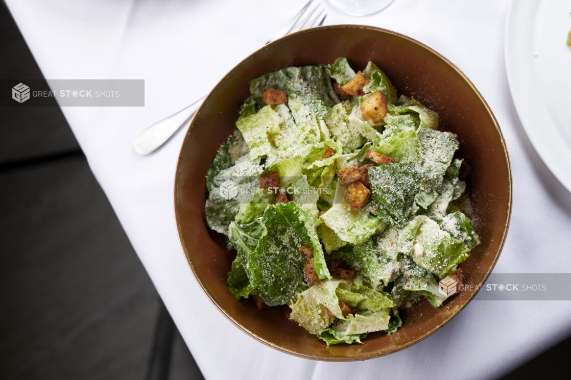 Caesar salad with croutons and parmesan cheese in a brown ceramic bowl on white linen with a fork on the side
