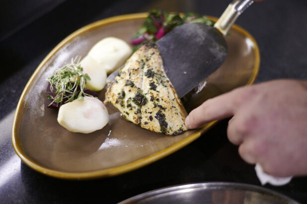 Pan seared fish on a brown ceramic plate with potatoes and micro greens garnish, hands plating the fish