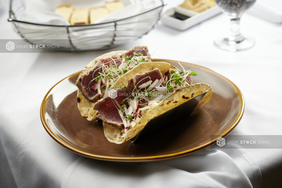 Seared tuna tacos with radish slaw and micro greens in metal taco holders on a rustic ceramic plate on a white linen background