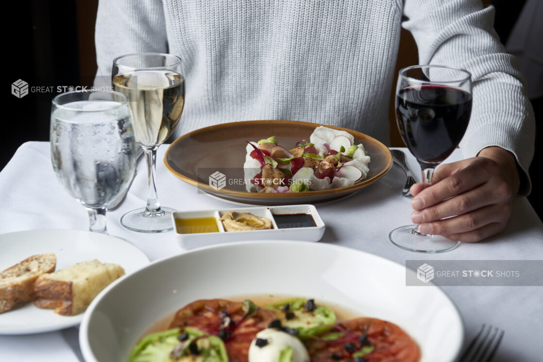 POV shot across a table from a man seated in front of a plate of food and holding the stem of a glass of red wine