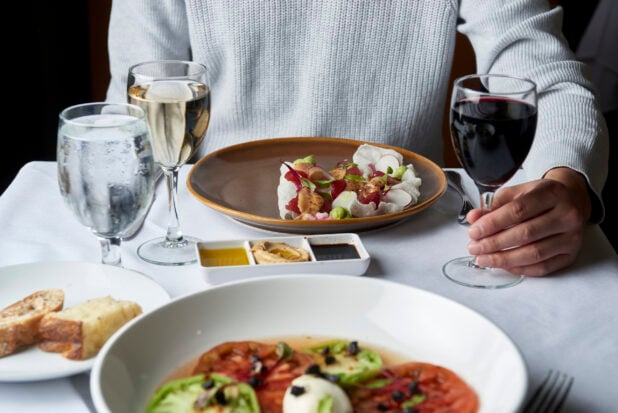 POV shot across a table from a man seated in front of a plate of food and holding the stem of a glass of red wine