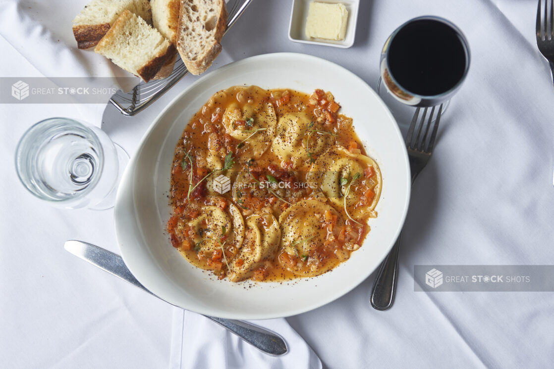 Ravioli with red sauce in a white angled bowl in a dining setting with bread, butter, a glass of  wine and a glass of water