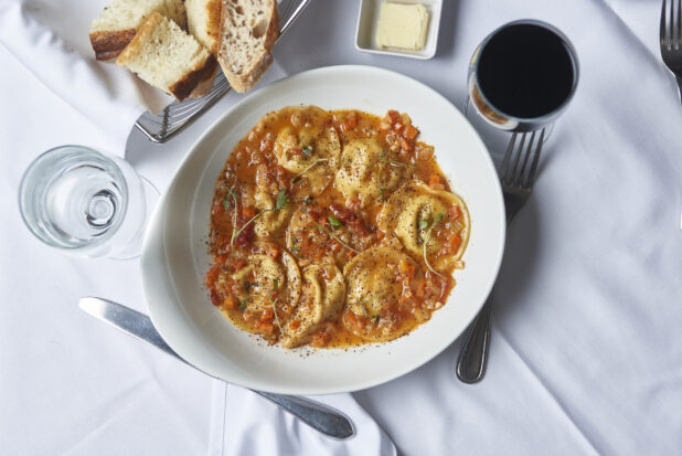 Ravioli with red sauce in a white angled bowl in a dining setting with bread, butter, a glass of  wine and a glass of water