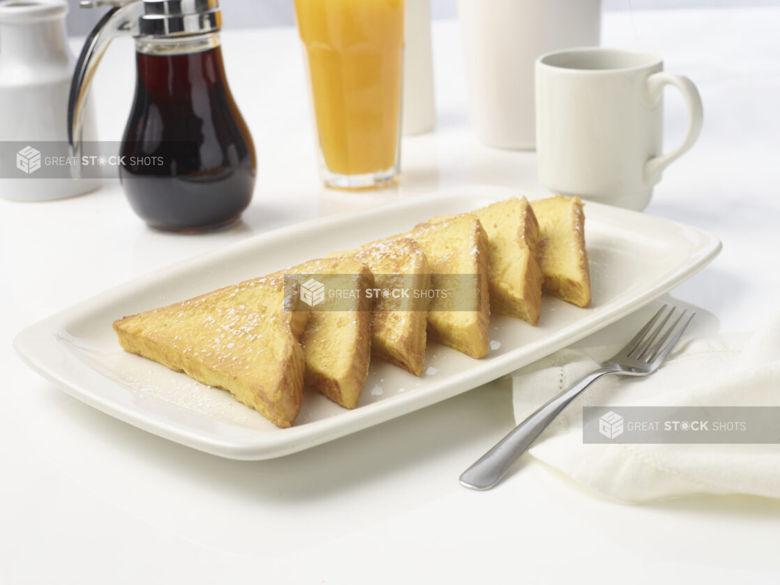 Plate of french toast dusted in icing sugar with orange juice, a coffee and syrup in the background on white