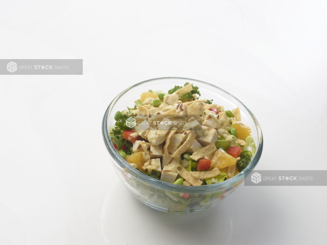 Chicken salad with kale and iceberg lettuce, red peppers, edamame and oranges drizzled with a creamy sauce in a glass bowl on a white background