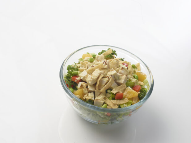 Chicken salad with kale and iceberg lettuce, red peppers, edamame and oranges drizzled with a creamy sauce in a glass bowl on a white background
