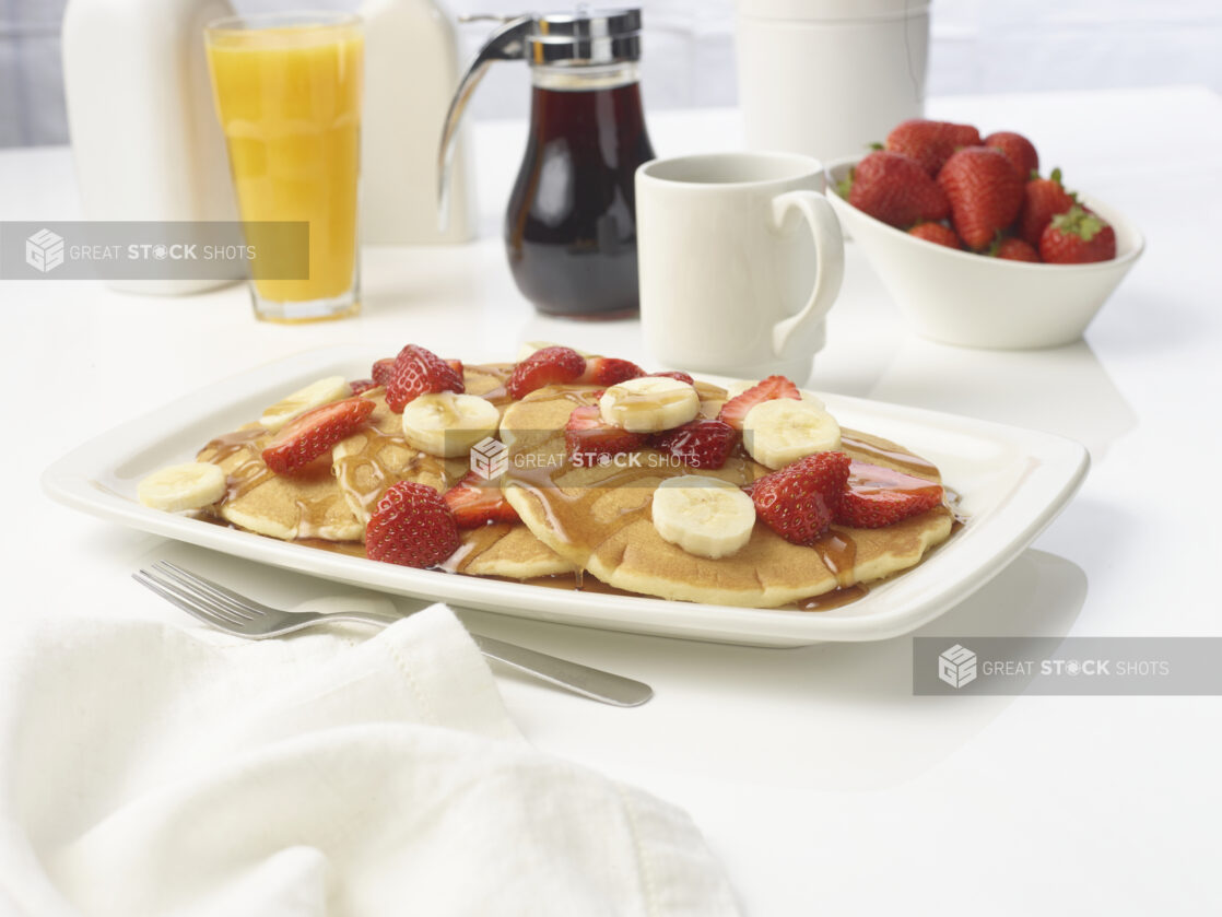 Breakfast plate of pancakes topped with fresh sliced strawberries and bananas and drizzled with syrup with a bowl of strawberries and drinks in the background