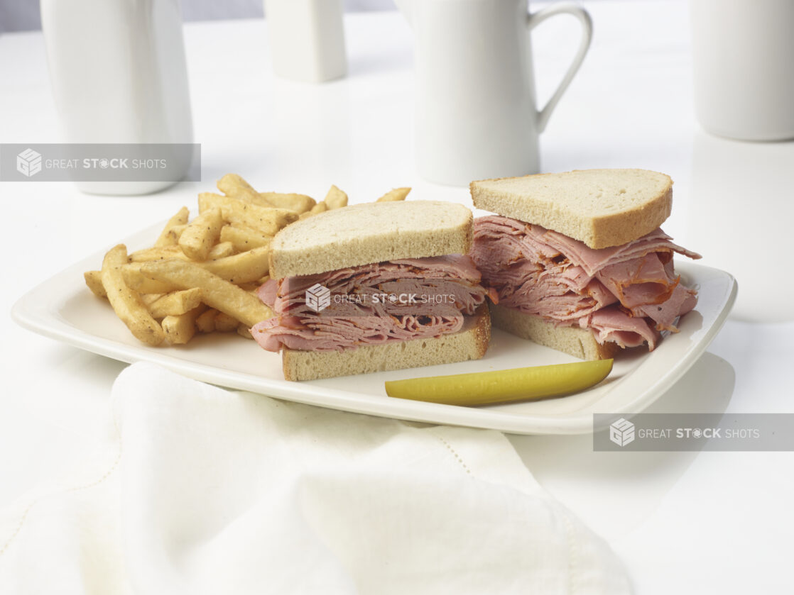 Corned beef sandwich on rye with a side of french fries and a dill pickle spear on a white plate on a white background