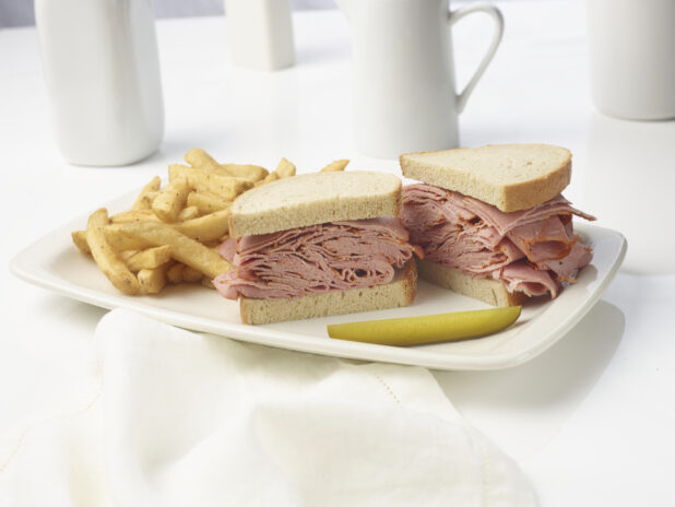 Corned beef sandwich on rye with a side of french fries and a dill pickle spear on a white plate on a white background