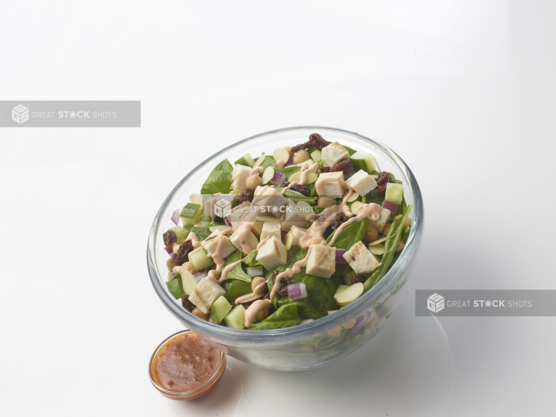 Spinach salad with grilled chicken, cucumber, chick peas, dried cranberries and sliced almonds with a creamy sauce drizzled on top in a glass bowl on a white background with a side vinaigrette