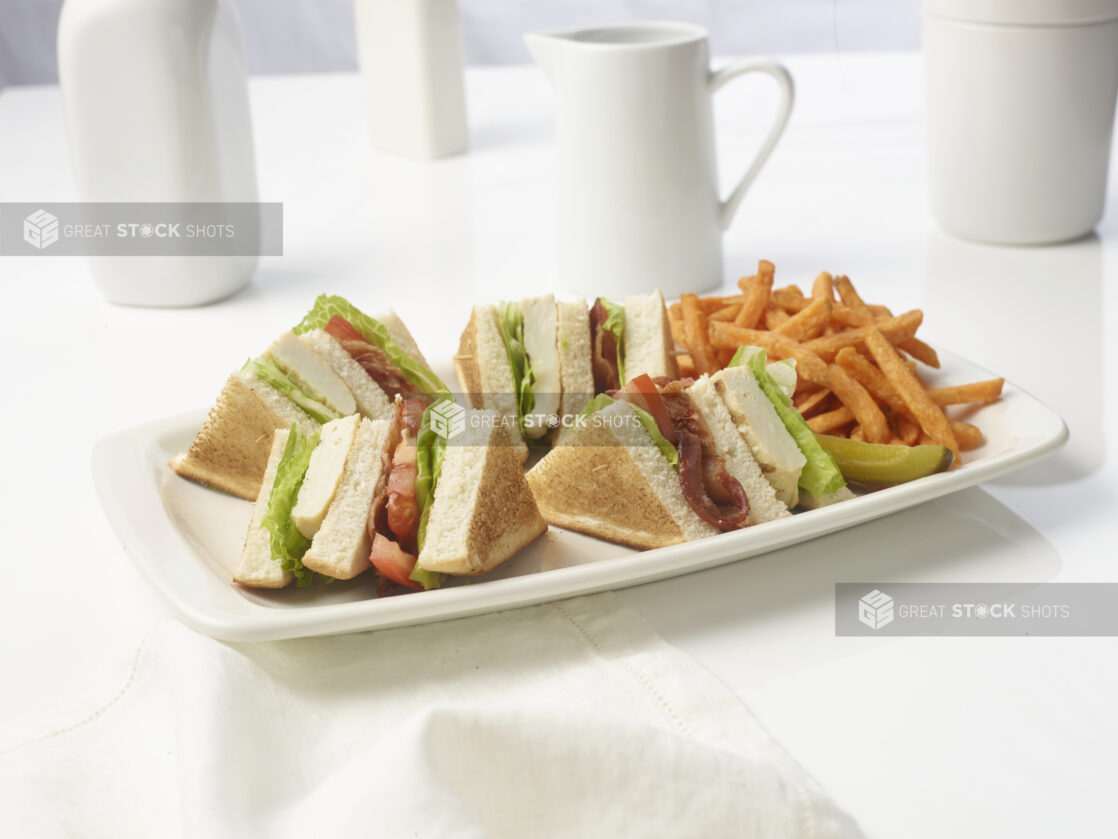 Quartered triple-decker chicken BLT on toasted white bread with a side of sweet potato fries and a dill pickle spear on a white plate