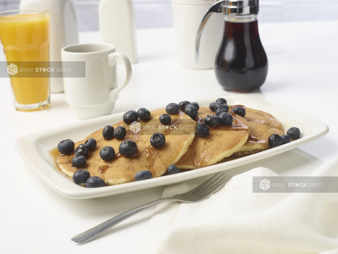 Breakfast plate of pancakes topped with fresh blueberries and drizzled syrup with a glass of orange juice and syrup in the background