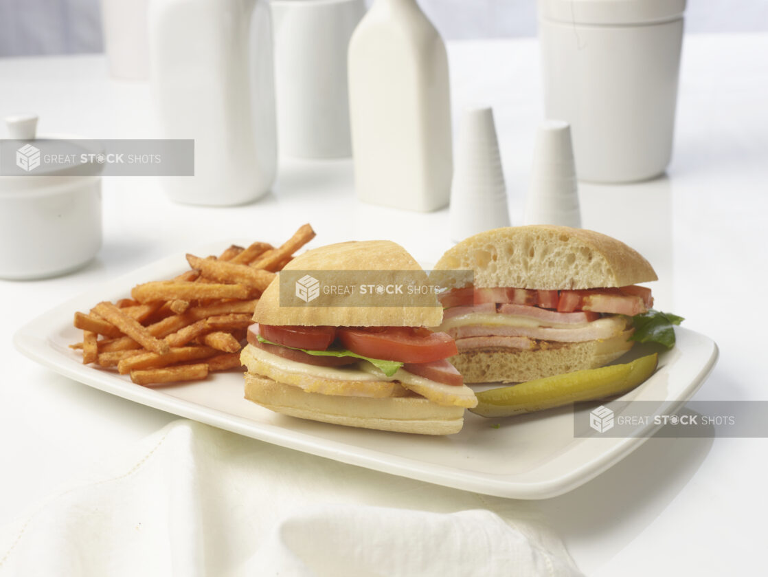 Breakfast sandwich with peameal bacon, sliced ham, melted cheese, tomato and lettuce on a white bun with a side of sweet potato fries and a pickle spear on a white plate