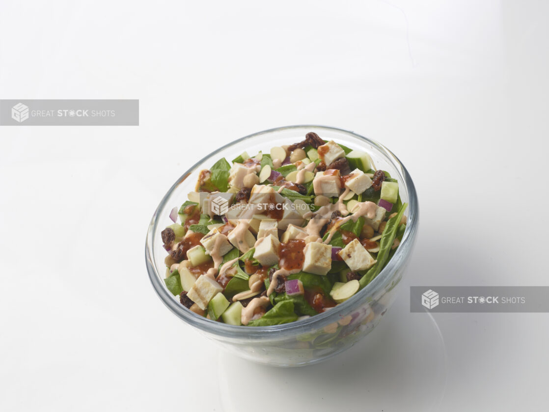 Spinach salad with grilled chicken, cucumber, chick peas, dried cranberries and sliced almonds with two different sauces drizzled on top in a glass bowl on a white background