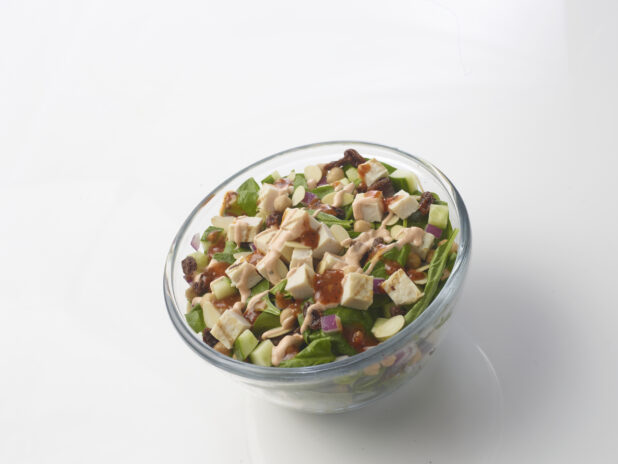 Spinach salad with grilled chicken, cucumber, chick peas, dried cranberries and sliced almonds with two different sauces drizzled on top in a glass bowl on a white background