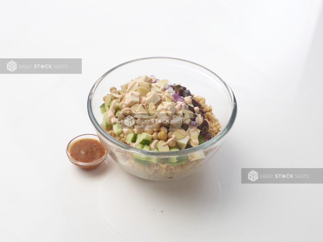 Grain salad in a glass bowl with chicken, cucumber, brown rice, chick peas, dried cranberries and sliced almonds, drizzled with a creamy sauce on a white background with a side of dressing