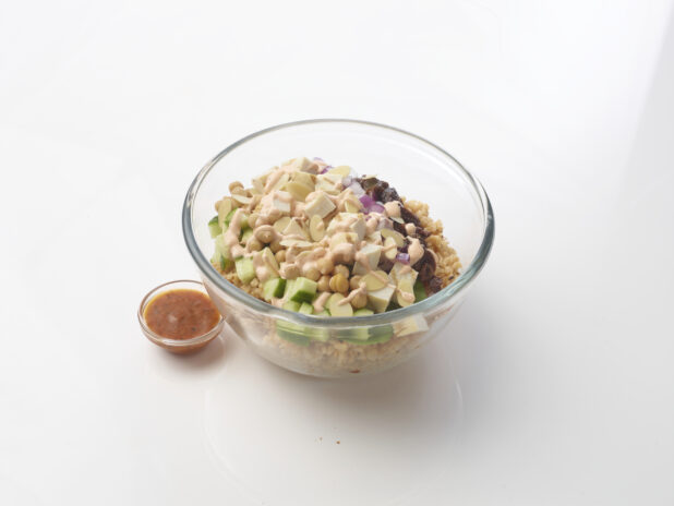 Grain salad in a glass bowl with chicken, cucumber, brown rice, chick peas, dried cranberries and sliced almonds, drizzled with a creamy sauce on a white background with a side of dressing