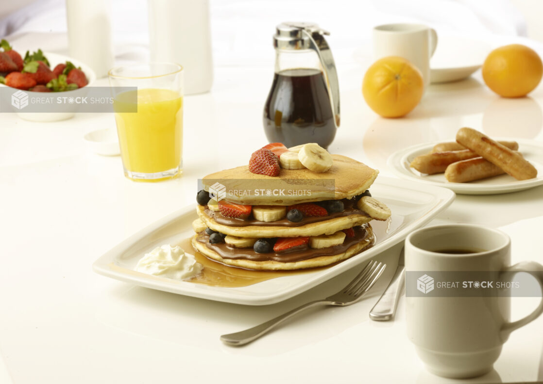 Pancakes layered with chocolate spread and sliced bananas, strawberries and blueberries with a side order of sausage and a cup of coffee on a white table