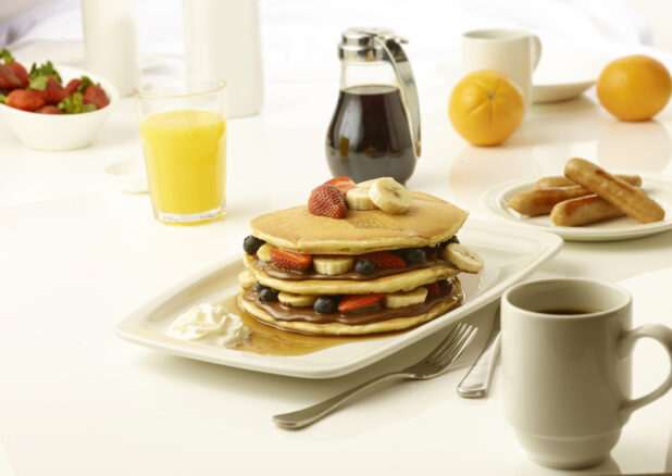 Pancakes layered with chocolate spread and sliced bananas, strawberries and blueberries with a side order of sausage and a cup of coffee on a white table