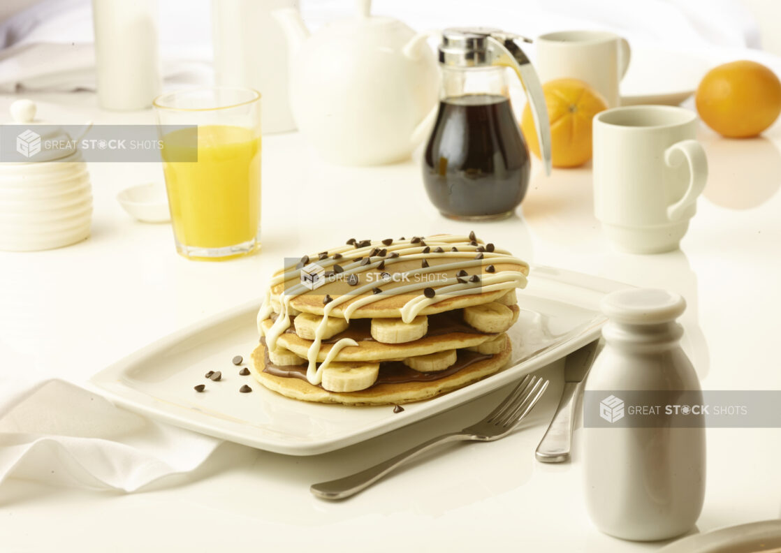 Pancakes layered with chcocolate spread and sliced bananas topped with chocolate chips and icing drizzle with a cup of coffee and orange juice in the background