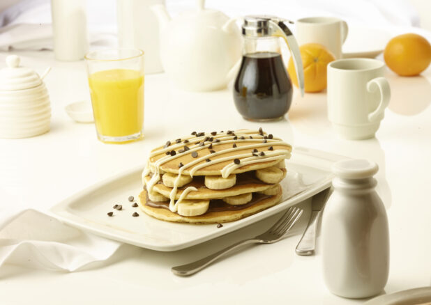 Pancakes layered with chcocolate spread and sliced bananas topped with chocolate chips and icing drizzle with a cup of coffee and orange juice in the background
