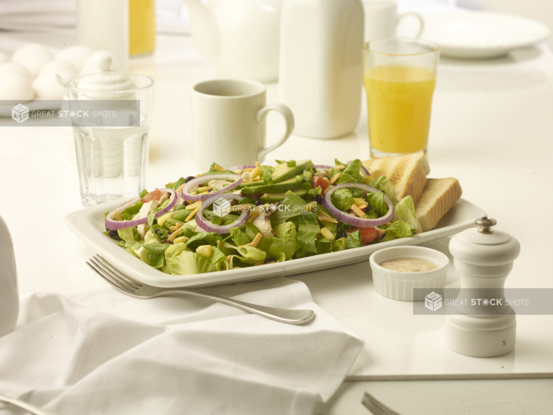 Large grilled chicken salad with side of toast and creamy dressing on a white plate with orange juice in the background on white