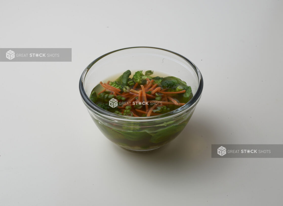 Small glass bowl of clear vegetable soup with broccoli, spinach, green onion and carrot on a white background