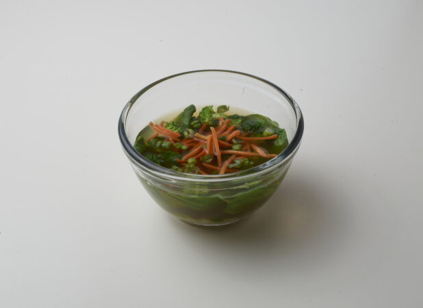 Small glass bowl of clear vegetable soup with broccoli, spinach, green onion and carrot on a white background