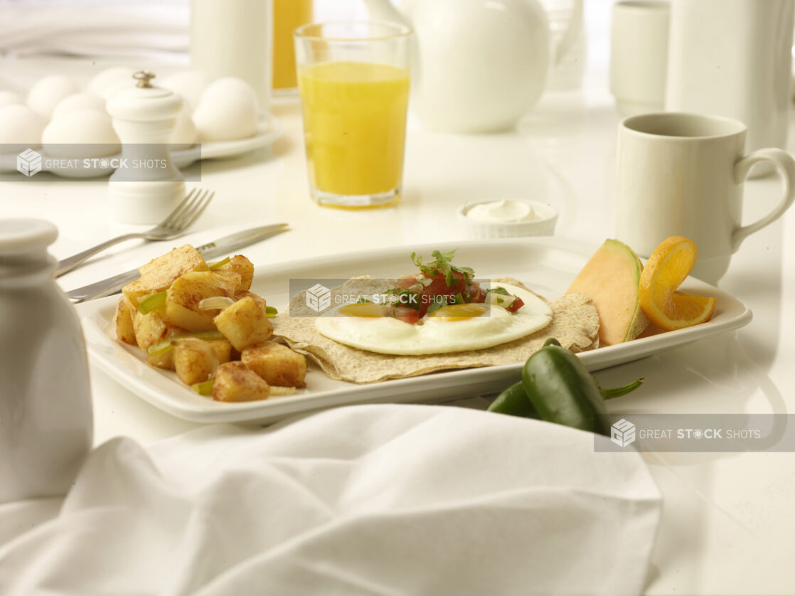 Breakfast of sunny side up eggs on stack of whole wheat wraps with side of home fries with a glass of orange juice and a cup of coffee in the background