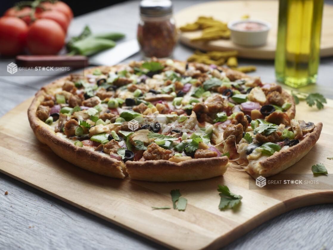 Sliced pizza with chicken, sausage, mushrooms, red onion and peppers topped with fresh cilantro and green onion on a wooden cutting board surrounded by ingredients in the background