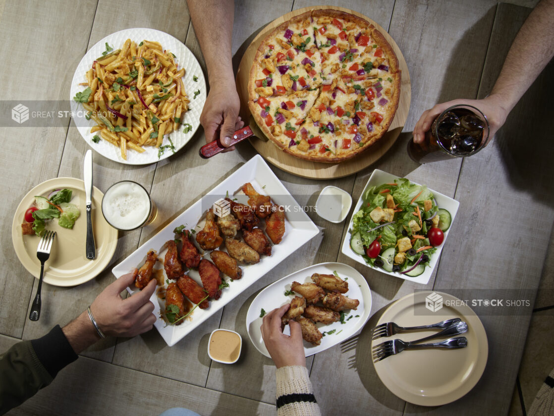 Overhead grouping of pizza, pasta, wings and a salad with hands in action on a wooden table