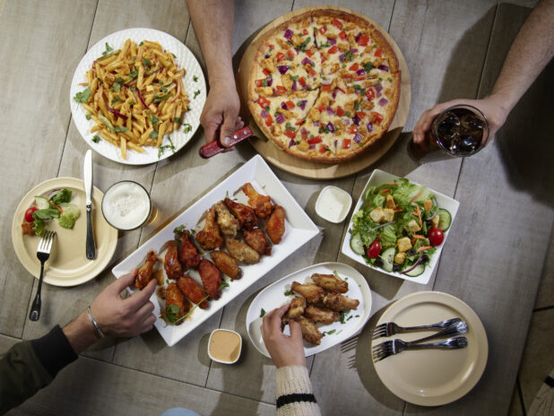 Overhead grouping of pizza, pasta, wings and a salad with hands in action on a wooden table