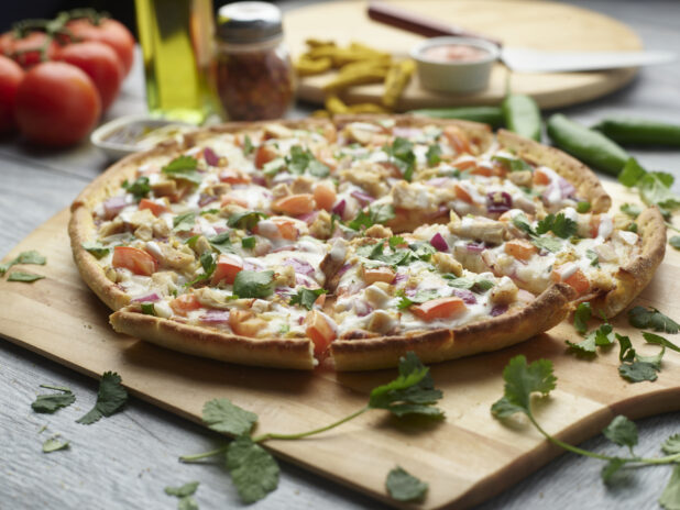 Sliced tandoori veggie pizza with mushrooms, red onion and tomatoes topped with fresh cilantro on a wooden cutting board surrounded by ingredients in the background
