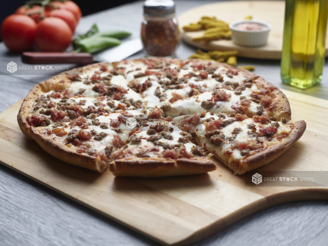 Sliced double meat pizza with ground beef and bacon on a wooden cutting board surrounded by ingredients in the background