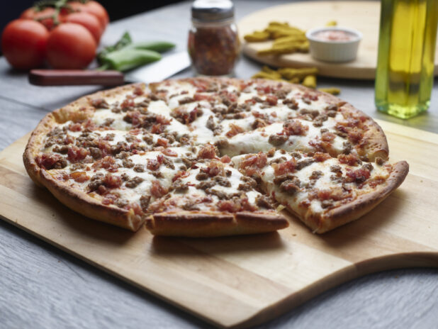 Sliced double meat pizza with ground beef and bacon on a wooden cutting board surrounded by ingredients in the background