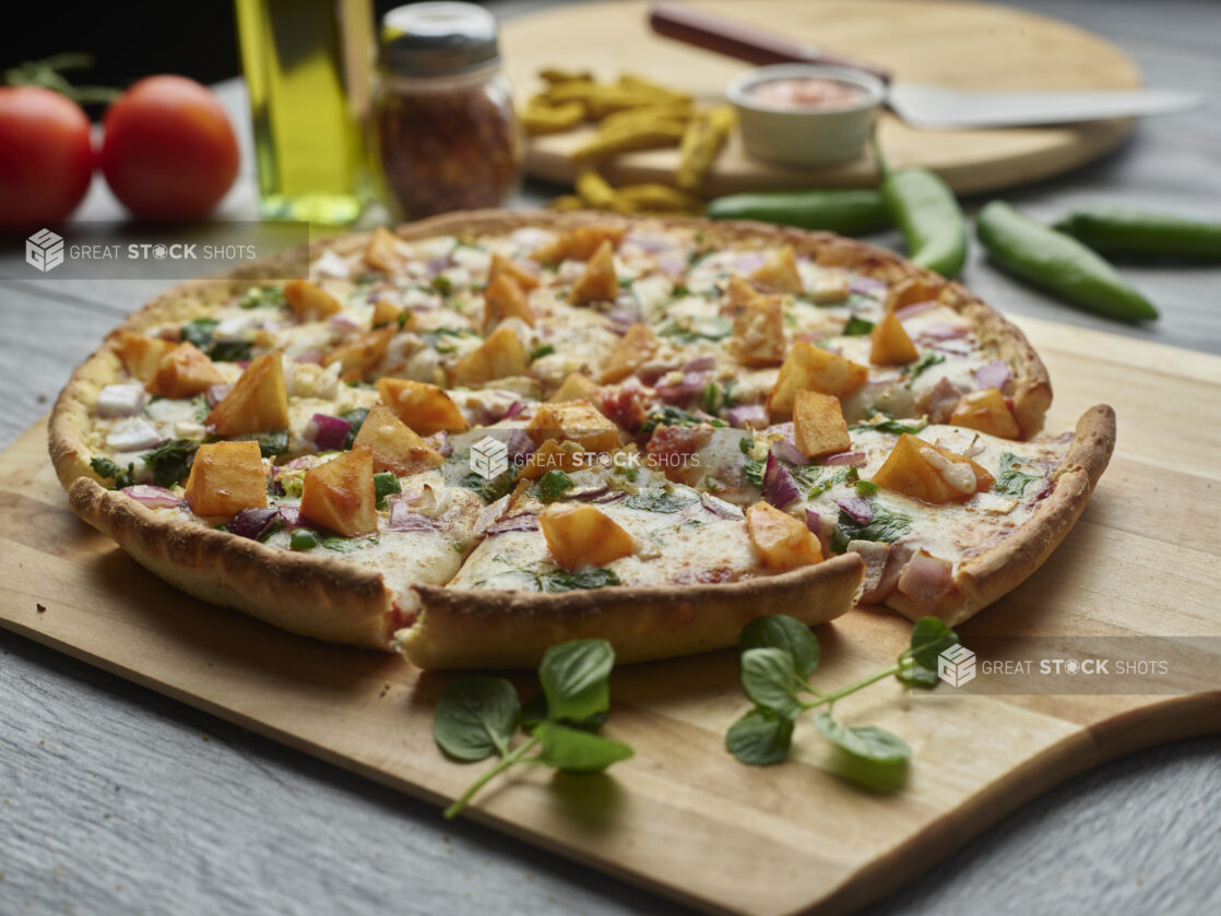 Sliced Aloo Chaat pizza with spiced potatoes, red onion and spinach on a wooden peel surrounded by ingredients