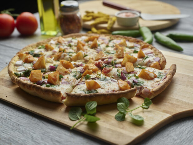 Sliced Aloo Chaat pizza with spiced potatoes, red onion and spinach on a wooden peel surrounded by ingredients