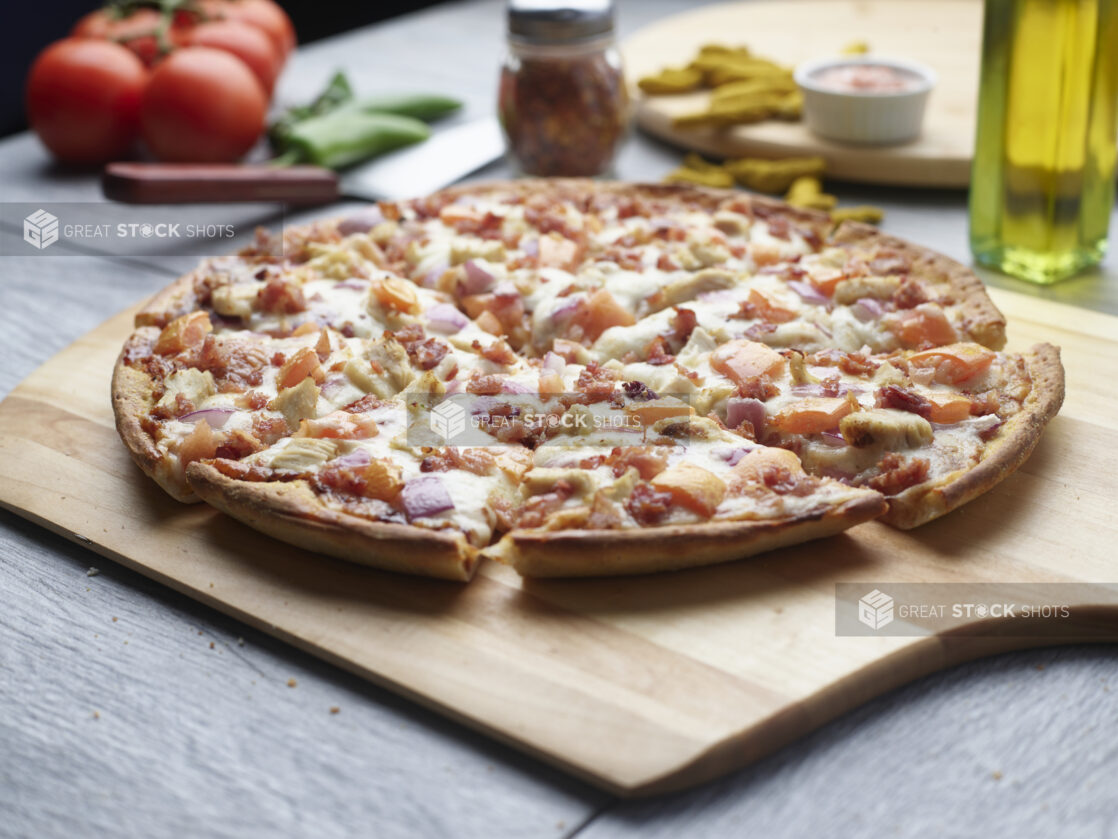 Sliced pizza with chicken, bacon, tomatoes and red onion on a wooden cutting board surrounded by ingredients in the background